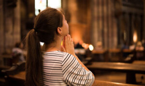 Religious young woman sitting on the bench in catholic church praying, looking up during church rule